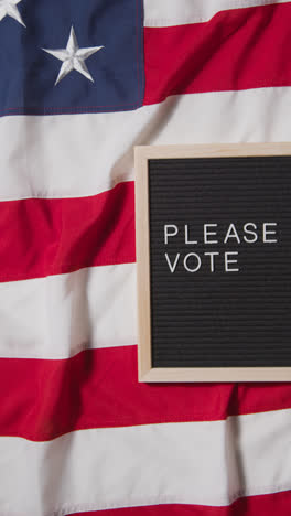 Vertical-Video-Of-Sign-Reading-Please-Vote-Lying-On-US-Stars-And-Stripes-Flag-For-American-Election-2024-2